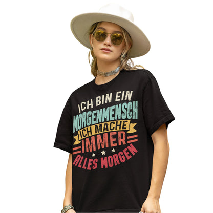 Frau trägt Schwarzes T-Shirt mit Spruch „Ich bin ein Morgenmensch – Ich mache immer alles morgen“ in bunten Retro-Farben