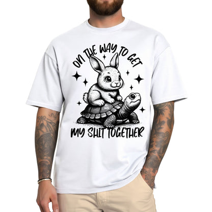 Weißes T-Shirt mit schwarzem Aufdruck: Ein Hase reitet auf einer Schildkröte mit dem Spruch „On the way to get my shit together“.