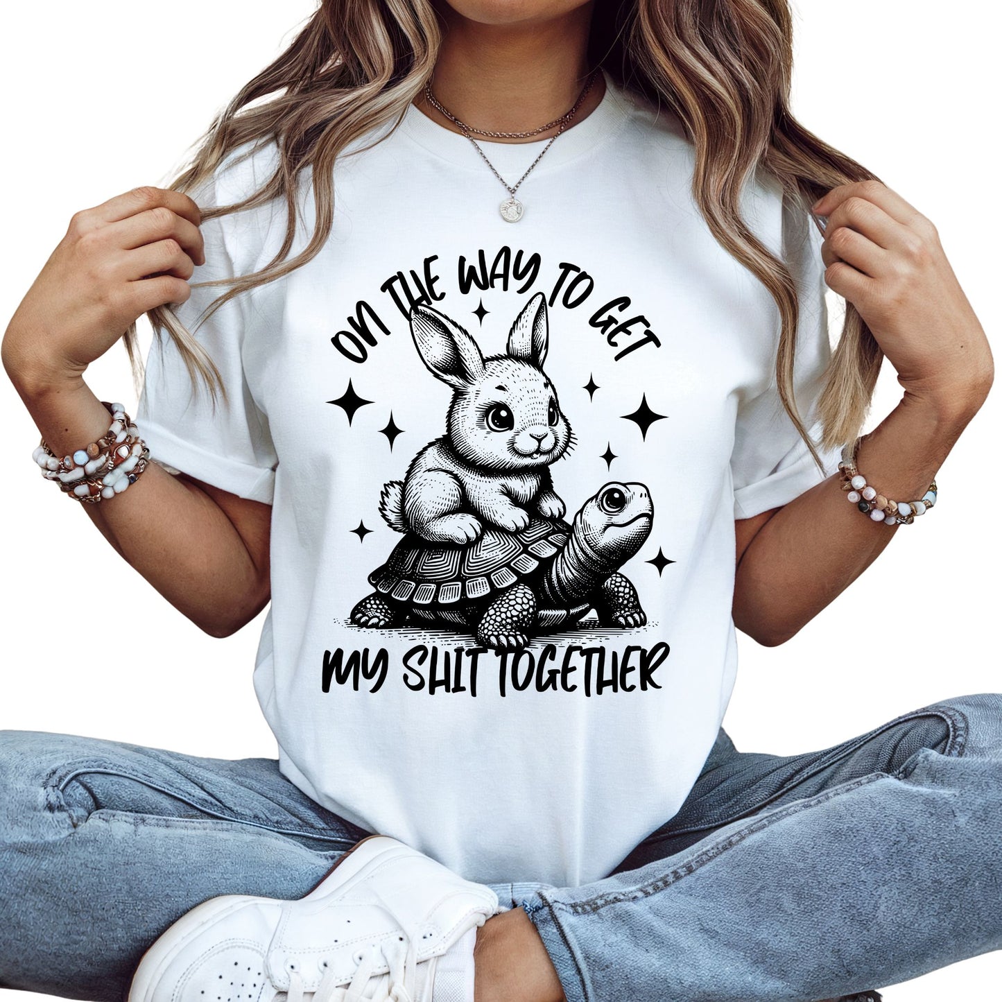 Weißes T-Shirt mit schwarzem Aufdruck: Ein Hase reitet auf einer Schildkröte mit dem Spruch „On the way to get my shit together“.
