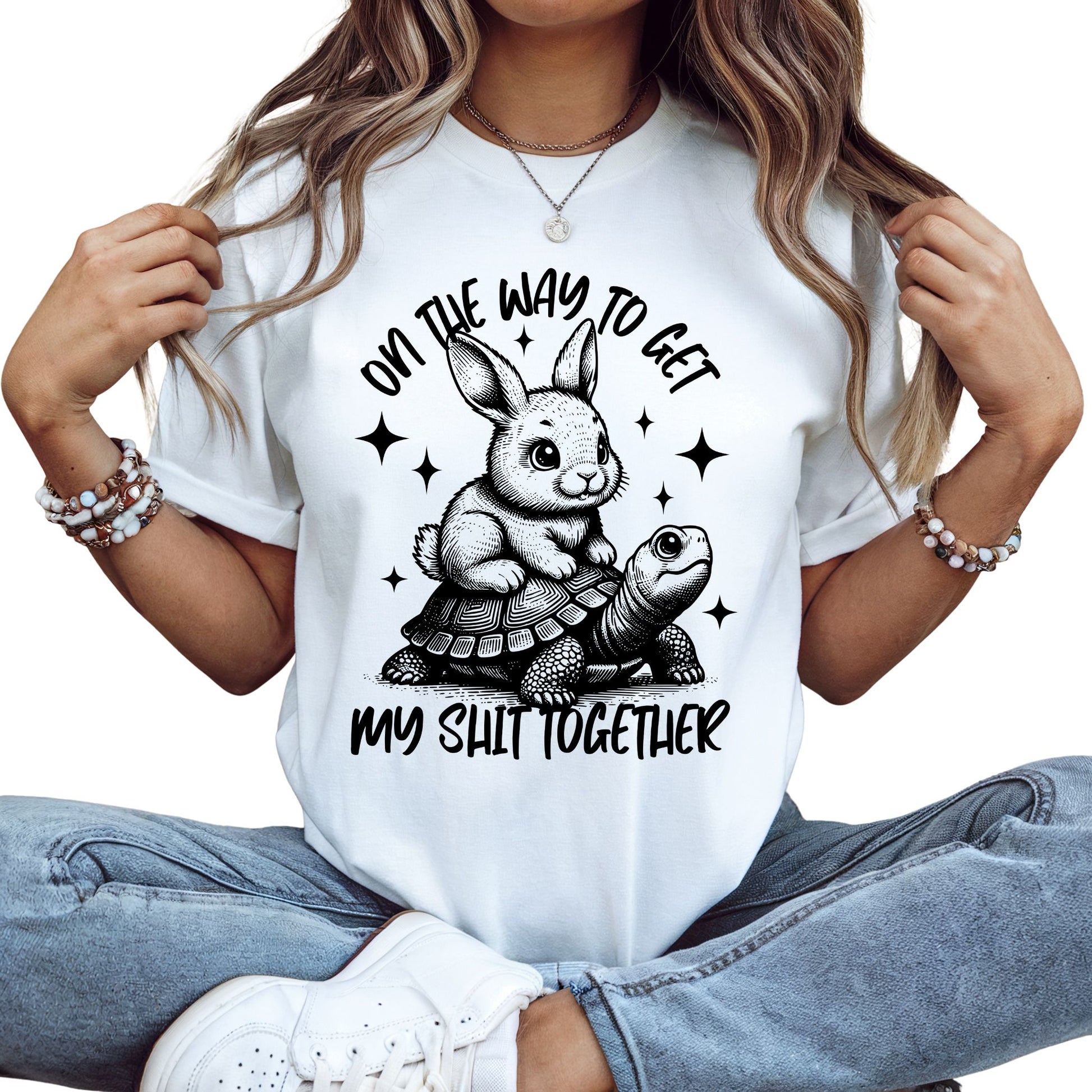 Weißes T-Shirt mit schwarzem Aufdruck: Ein Hase reitet auf einer Schildkröte mit dem Spruch „On the way to get my shit together“.
