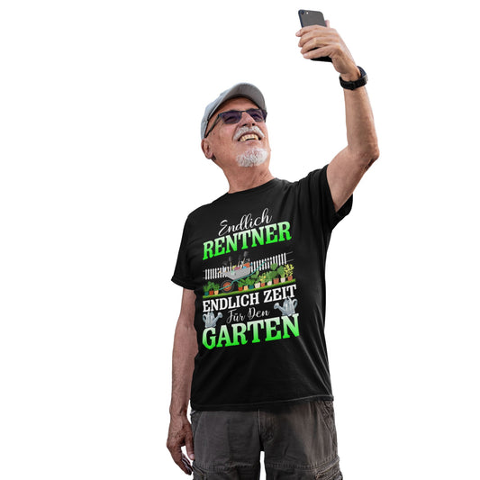 Älterer Mann mit schwarzem T-Shirt „Endlich Rentner – Endlich Zeit für den Garten“, lachend beim Selfie, Gartenmotiv mit Rasenmäher und Pflanzen