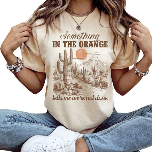 T-Shirt mit Spruch 'Something in the Orange tells me we’re not done' und Wüstenmotiv mit Kaktus & Sonnenuntergang – emotionales Country Shirt