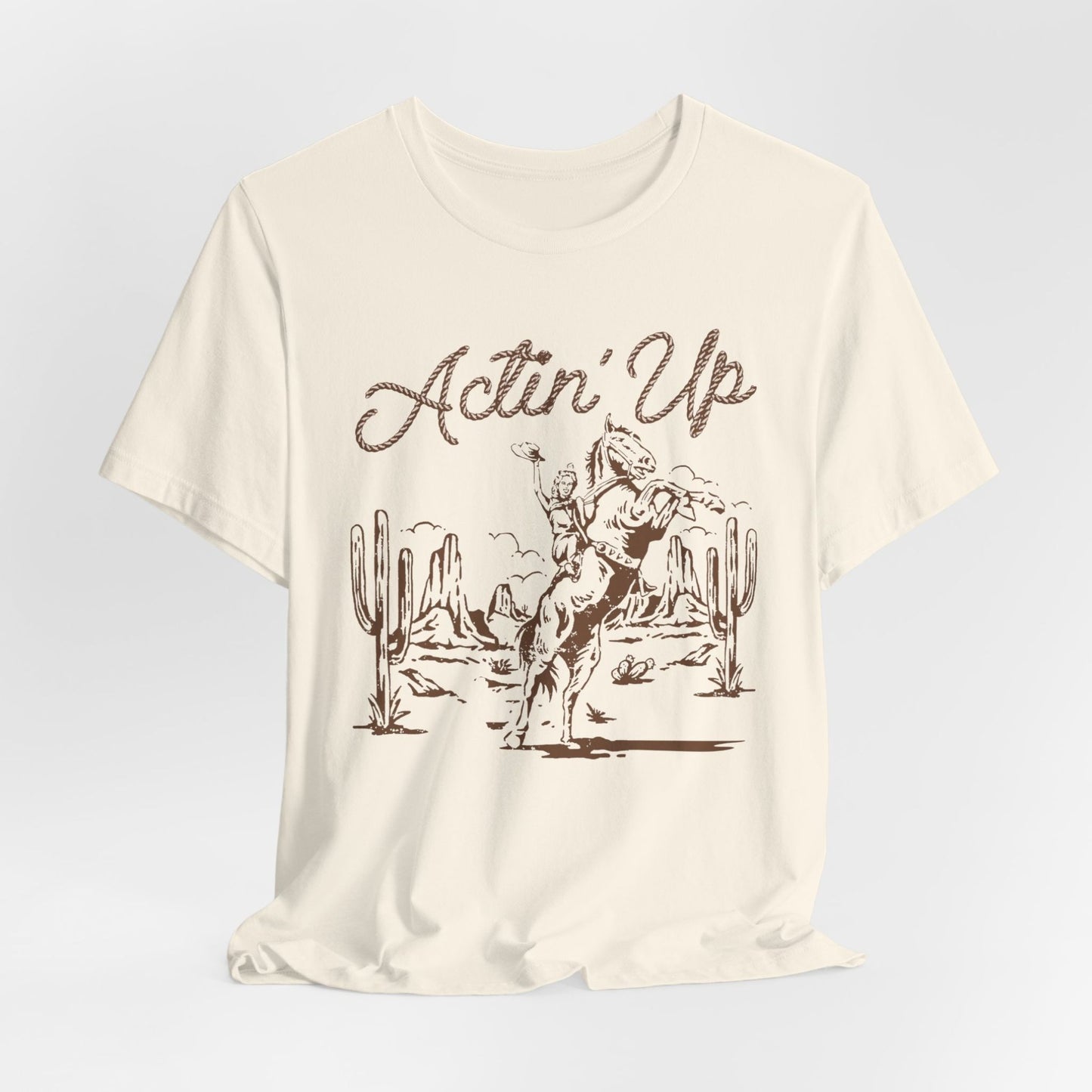 T-Shirt mit 'Actin Up' in Lasso-Schrift und Illustration einer reitenden Frau auf Pferd in der Wüste – Western-Style Design