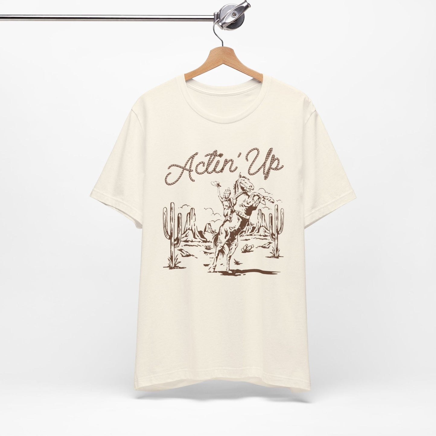T-Shirt mit 'Actin Up' in Lasso-Schrift und Illustration einer reitenden Frau auf Pferd in der Wüste – Western-Style Design