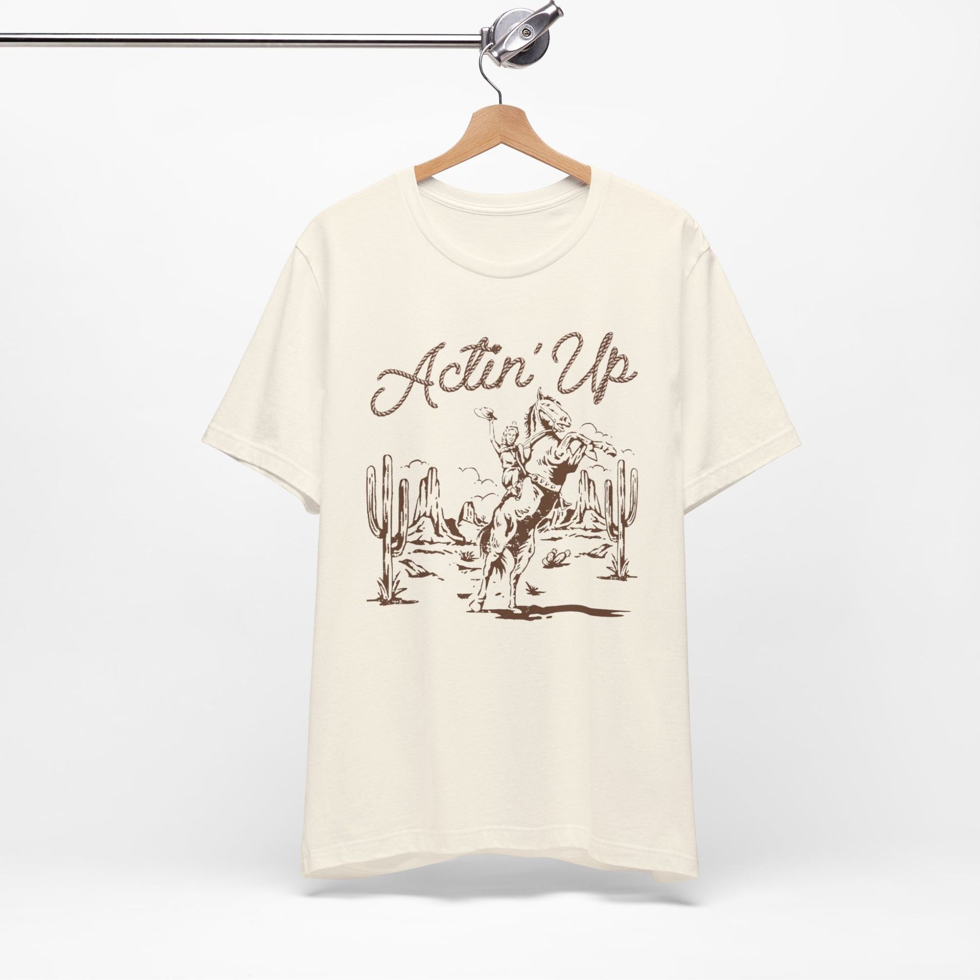 T-Shirt mit 'Actin Up' in Lasso-Schrift und Illustration einer reitenden Frau auf Pferd in der Wüste – Western-Style Design