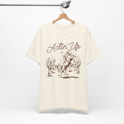 T-Shirt mit 'Actin Up' in Lasso-Schrift und Illustration einer reitenden Frau auf Pferd in der Wüste – Western-Style Design