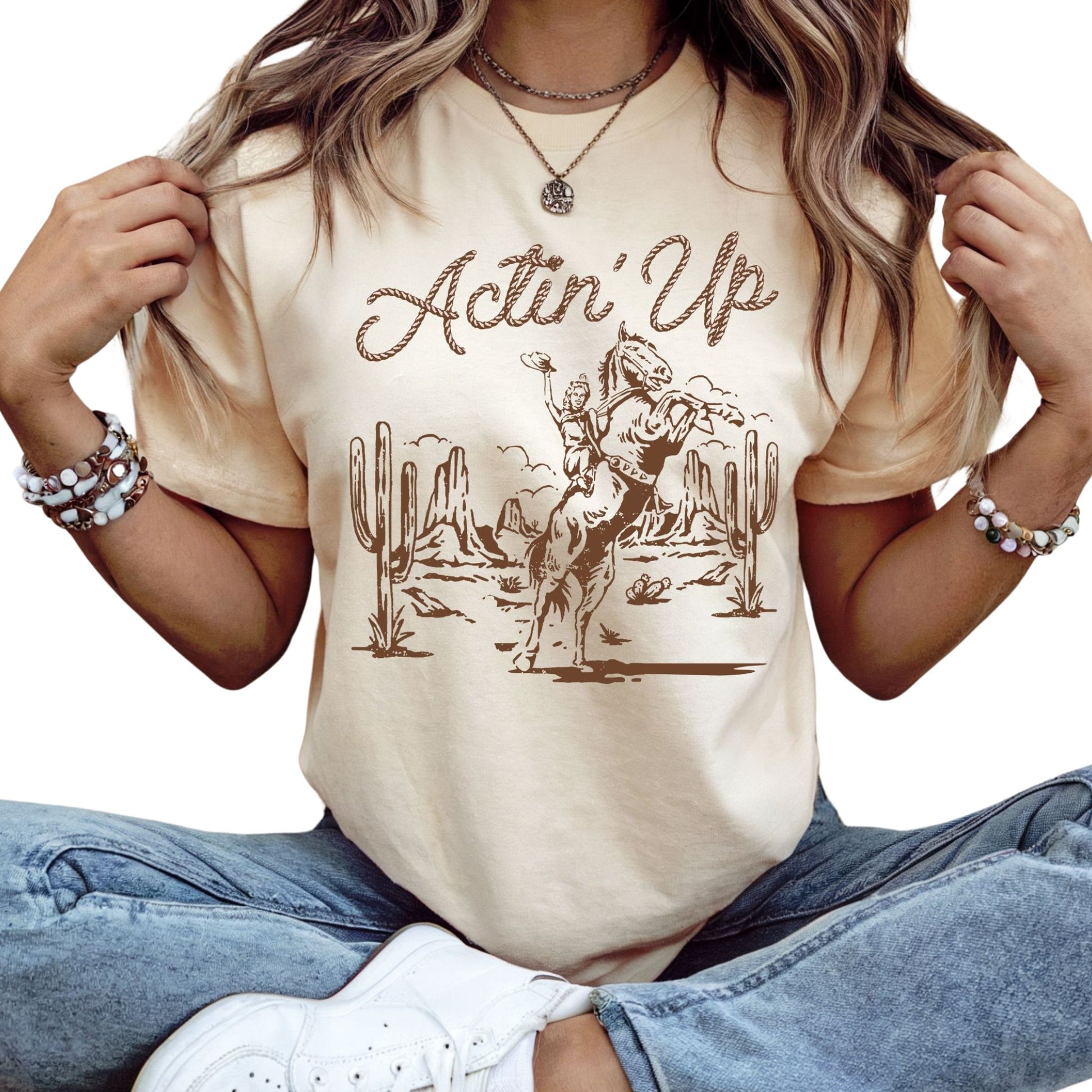 T-Shirt mit 'Actin Up' in Lasso-Schrift und Illustration einer reitenden Frau auf Pferd in der Wüste – Western-Style Design