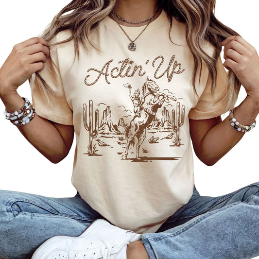 T-Shirt mit 'Actin Up' in Lasso-Schrift und Illustration einer reitenden Frau auf Pferd in der Wüste – Western-Style Design